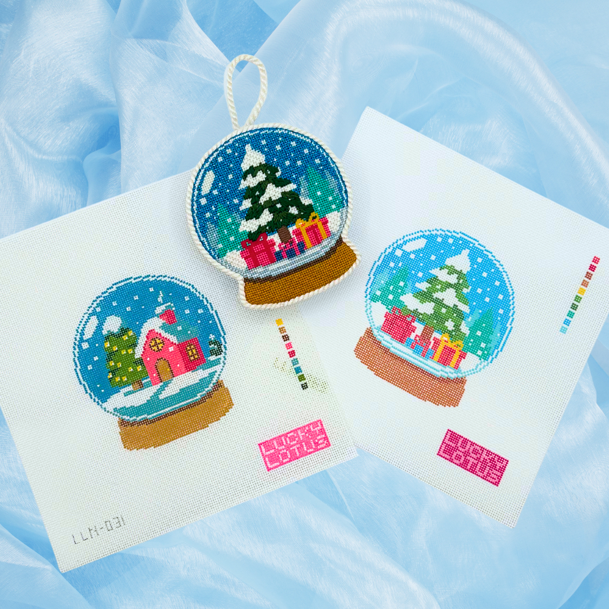 Lucky Lotus Needlepoint Christmas snow globe designs featuring a snowy Christmas tree with colorful presents and a pink winter village, displayed as two needlepoint canvases and a finished ornament against a soft blue background.