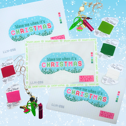 A flat-lay product display of a Christmas-themed needlepoint canvas kit by Lucky Lotus, featuring a central white canvas with the design 'Wake me when it's CHRISTMAS' in pastel blue, pink, and green, surrounded by coordinating craft supplies including thread spools, keychains, and small accessories on a light blue snowflake-patterned background.