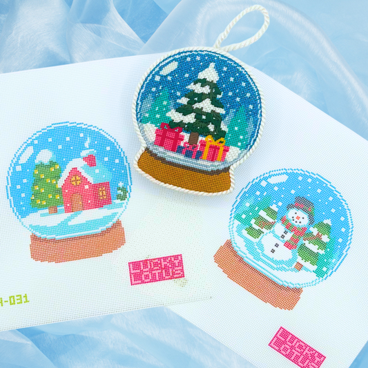 Lucky Lotus Needlepoint Snow Globe Collection featuring the 2024 pink Christmas house canvas, finished 2025 Christmas tree and presents ornament, and 2026 snowman canvas displayed on light blue fabric.