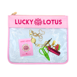 A Lucky Lotus branded clear zippered pouch with a pink top border and logo, shown on a white background. Inside the pouch are several small accessories, including a gold keychain, a pink cylindrical item, green scissors, and a pink card.