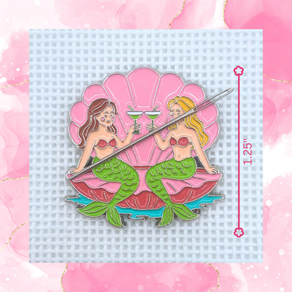 Close-up of the Mermaids Needle Minder by Lucky Lotus Needlepoint featuring two mermaids sitting in a pink seashell while toasting with martinis, displayed on needlepoint canvas with a needle attached.