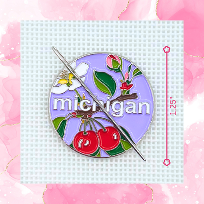 Michigan state needlepoint needle minder close up featuring cherries, apple blossom, greenery, and a needle on a purple background.