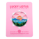 North Carolina needle minder featuring layered blue mountains, evergreen trees, a colorful sunset, and North Carolina lettering.