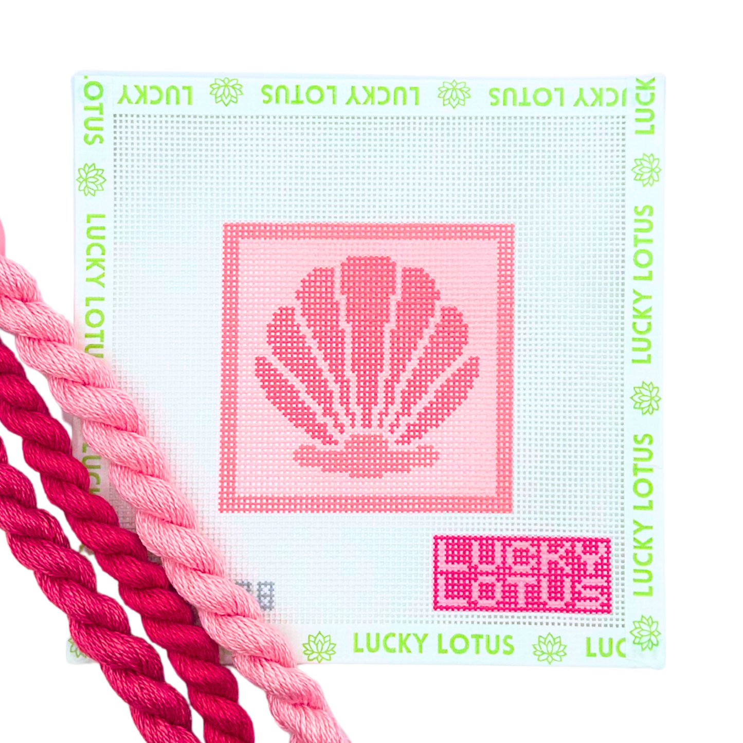 A Lucky Lotus needlepoint canvas featuring a pink seashell design in the center, surrounded by a border of green 'LUCKY LOTUS' text and flower motifs. The canvas is shown with two pink needlepoint thread skeins in the foreground on a clean white background.