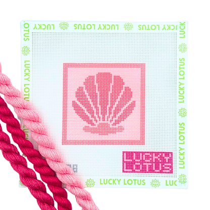 A Lucky Lotus needlepoint canvas featuring a pink seashell design in the center, surrounded by a border of green 'LUCKY LOTUS' text and flower motifs. The canvas is shown with two pink needlepoint thread skeins in the foreground on a clean white background.