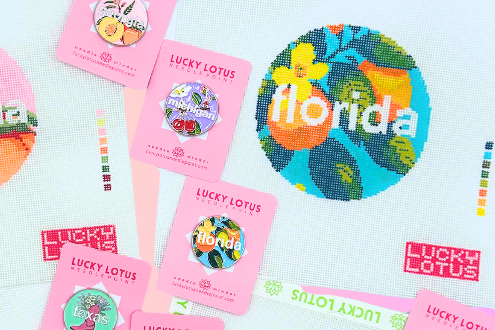 A flat lay of multiple pink Lucky Lotus sticker sheets and circular embroidered-style state-themed patches on a pastel gradient background. Visible designs include Georgia, Florida, Hawaii, Tennessee, and North Carolina, with colorful fruit, guitar, and landscape motifs.